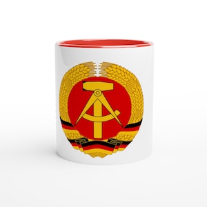 Könnte beinhalten: Weißer Keramikbecher mit rotem Innenraum. Der Becher zeigt ein rundes Emblem mit gelbem Hammer und Zirkel auf rotem Hintergrund, umgeben von Weizenähren und einem schwarz-rot-goldenen Band.