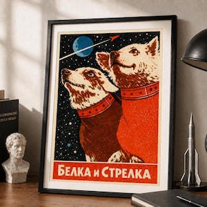 May include: A framed poster featuring a space-themed illustration of two dogs wearing red collars against a starry night sky with a blue moon and a rocket. The text "БЕЛКА и СТРЕЛКА" is printed in white at the bottom on a red rectangular background.