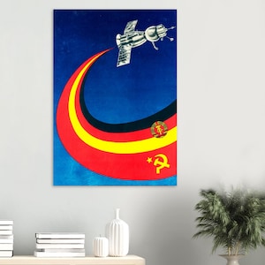 May include: Vintage space-themed poster with a satellite and a curved trail of red, yellow, and black. The poster features a blue background and a communist symbol. The artwork is mounted on a white wall.