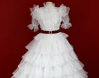 May include: A white, tiered formal dress with a high collar and ruffled sleeves. The dress features a fitted bodice with small buttons and a burgundy belt with a silver buckle. The skirt is voluminous, with multiple layers of sheer fabric.