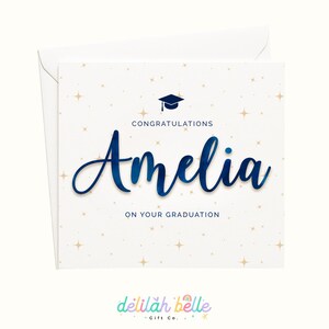 May include: A white graduation card with a matching envelope. The card features the name "Amelia" in a dark blue script, with the words "Congratulations" and "On Your Graduation" in a smaller font. Gold star accents are scattered across the card.