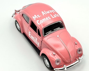 May include: A pink toy car with the text "Ms. Always Comes Late" on the roof and "Emily" on the side. The car has a classic design with chrome accents and black tires.