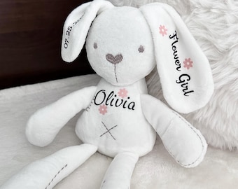 May include: White plush bunny rabbit toy with embroidered details. The bunny has a pink heart nose, pink flower accents, and the name "Olivia" embroidered on its chest. The bunny's ear is embroidered with "Flower Girl".
