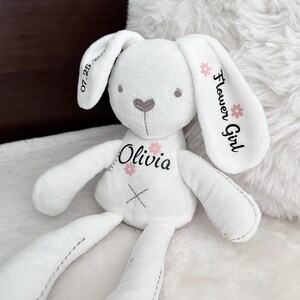 May include: White plush bunny rabbit toy with embroidered details. The bunny has a pink heart nose, pink flower accents, and the name "Olivia" embroidered on its chest. The bunny's ear is embroidered with "Flower Girl".