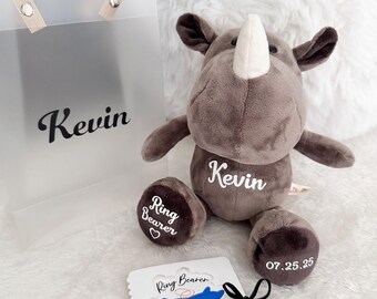 May include: A gray plush rhinoceros toy with the name "Kevin" embroidered on its chest. The toy is holding a white card with the text "Ring Bearer" and a blue rhinoceros illustration. The card is tied with a black ribbon. The toy is in a clear plastic bag with the name "Kevin" printed on it.