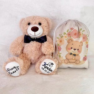 May include: A plush, light brown teddy bear wearing a black bow tie, with "Rings Bearer" and "Kevin 08.23.27" printed on its paws. A matching drawstring bag with a floral design and a cartoon bear holding a ring is also present.