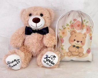 May include: A plush, light brown teddy bear wearing a black bow tie, with "Rings Bearer" and "Kevin 08.23.27" printed on its paws. A matching drawstring bag with a floral design and a cartoon bear holding a ring is also present.