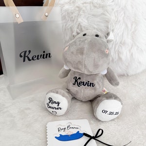 May include: A gray plush hippopotamus with the name "Kevin" embroidered on its chest. The hippo is holding a white card with the text "Ring Bearer" and a blue illustration of a ring. The hippo is sitting on a white fluffy surface.