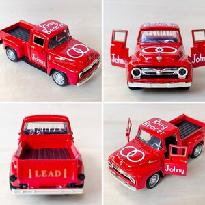May include: Four red toy pickup trucks with white lettering. The trucks have the words "Ring Bearer" and "Johny" on them. One truck has the words "Lead" on the back. The trucks have white rings on the sides.