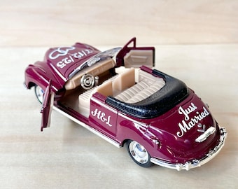 May include: A burgundy toy car with a black convertible top. The car has white lettering that says "Just Married" and "H&L" on the back and side. The car has a white ring design on the hood and the doors are open.