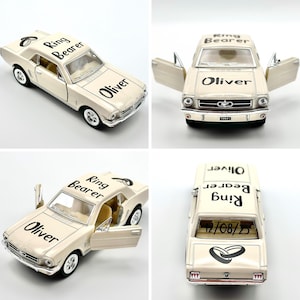 May include: A white toy car with black lettering that says "Ring Bearer" and "Oliver". The car has a black ring design on the roof and a date on the back window.