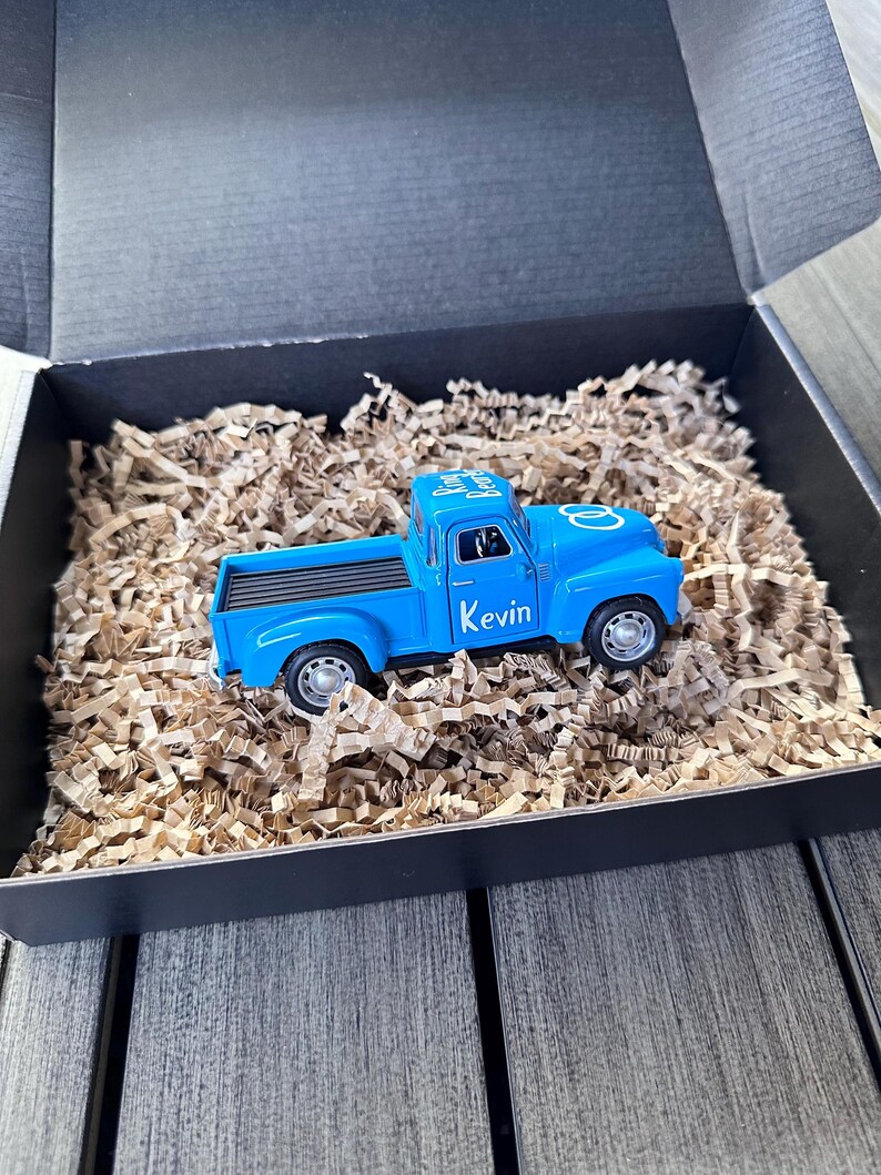 May include: A blue toy pickup truck with the words "Ring Bearer" and "Kevin" on the side. The truck is in a black gift box with brown shredded paper.