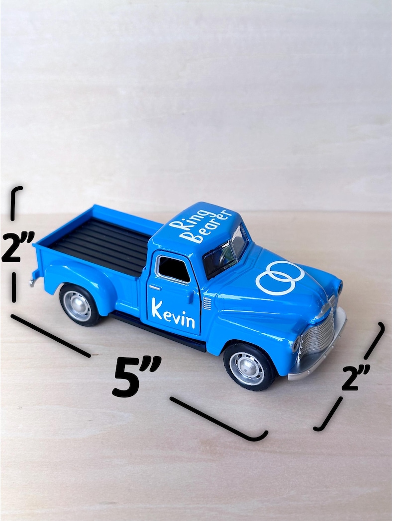 May include: A blue toy pickup truck with a white inscription that reads "Ring Bearer Kevin" and a white infinity symbol. The truck measures 5 inches long and 2 inches wide.