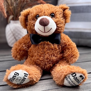 May include: A brown teddy bear with a black bow tie. The bear has a white muzzle and a brown nose. The bear's paws have text that reads "Ring Guard" and "Daniel Special Agent 05/26/2023".