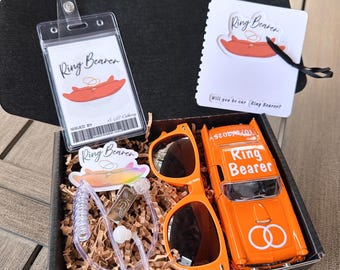 May include: A gift box with wedding-themed items. Includes an orange toy car, sunglasses, a badge holder, and cards, all with "Ring Bearer" text. The box is filled with brown paper and sits on a wooden surface.