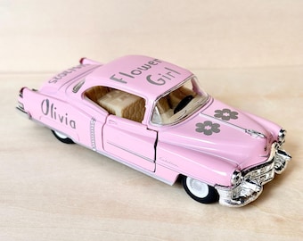 May include: A pink toy car with the words "Flower Girl" and "Olivia" on the side. The car has a silver grill and chrome accents.