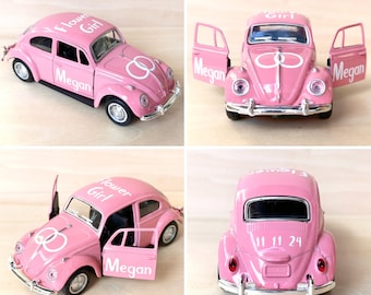 May include: A pink toy car with the words "Flower Girl" and "Megan" on the side. The car has a white ring design on the hood and the doors are open. The car has a white license plate with the numbers "11 11 24" on the back.