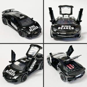 May include: A black toy sports car with white text reading RING BEARER on the hood, KEVIN on the side, and 2026 on the roof. The vehicle features scissor doors that open upward and a rear engine compartment that opens to reveal engine details.
