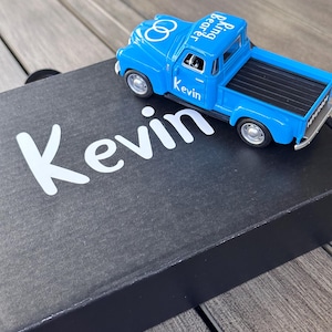 May include: A blue toy pickup truck with the words "Kevin" and "Bearing Rings" printed on the side. The truck is sitting on a black box with the word "Kevin" printed in white.