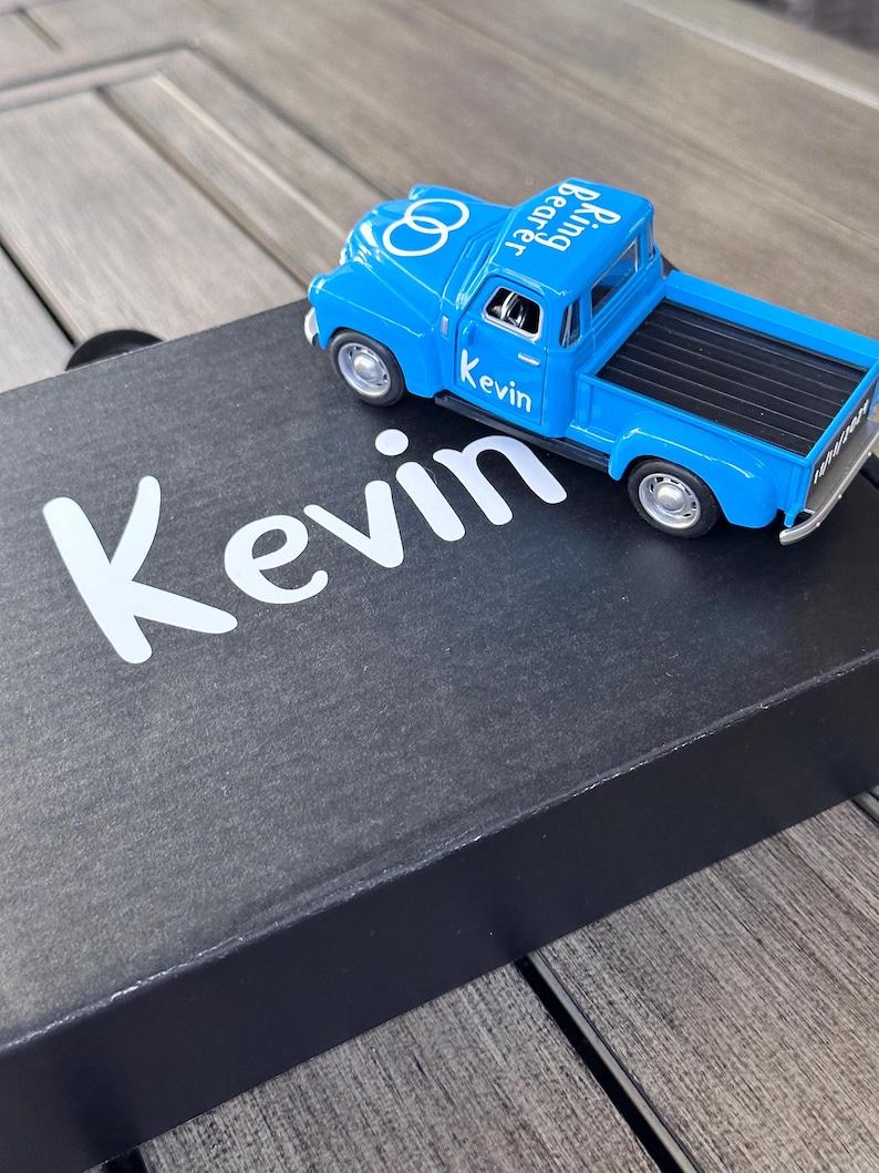 May include: A blue toy pickup truck with the words "Kevin" and "Bearing Rings" printed on the side. The truck is sitting on a black box with the word "Kevin" printed in white.