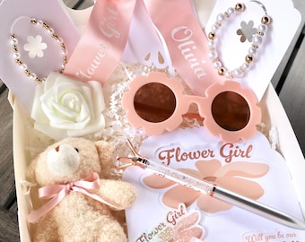 May include: A pink gift box filled with items for a flower girl. The box includes a teddy bear, sunglasses, a pen, a bracelet, a flower, and a sash that says "Flower Girl" and "Olivia".