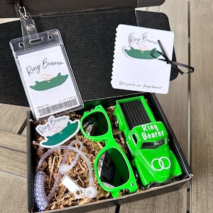 May include: A black gift box filled with ring bearer accessories. Includes a green toy truck, sunglasses, a badge holder, a sticker, and a card. The truck and card have "Ring Bearer" text. The box sits on a wooden surface.