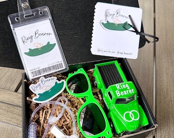 May include: A black gift box filled with ring bearer accessories. Includes a green toy truck, sunglasses, a badge holder, a sticker, and a card. The truck and card have "Ring Bearer" text. The box sits on a wooden surface.