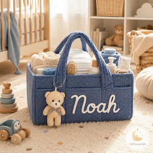 Könnte beinhalten: Ein rechteckiger Aufbewahrungskorb aus blauem Sherpa-Stoff mit zwei Griffen, auf dessen Vorderseite der Name Noah in weißer Schreibschrift gestickt ist. Ein kleiner beiger Plüschbär mit einer braun-weiß karierten Schleife ist am vorderen Griff befestigt.