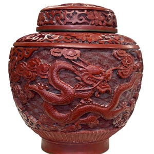 Op de afbeelding: Een rode pot met deksel en gedetailleerde gravures. De pot heeft een drakenontwerp, wolken en bloemmotieven. Het oppervlak heeft een textuur en het deksel is versierd met gebeeldhouwde bloemen.