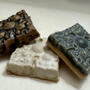 May include: Three ceramic tiles with intricate raised designs. The tiles are square and have a textured surface. One tile is brown with black accents, one is white with brown accents, and one is green with black accents.
