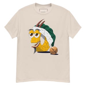 Peut inclure: T-shirt beige clair avec une illustration de dessin animé d'une créature jaune avec des yeux bruns, des cornes et une barbe. La créature porte un chapeau vert et blanc et a une cloche à côté. Le t-shirt a un col rond.