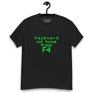Könnte beinhalten: Schwarzes T-Shirt mit dem Text "Keyboard not found. Press F4" in grüner Pixel-Schrift. Das T-Shirt hängt an einem schwarzen Kleiderbügel. Das T-Shirt hat einen Rundhalsausschnitt und kurze Ärmel.