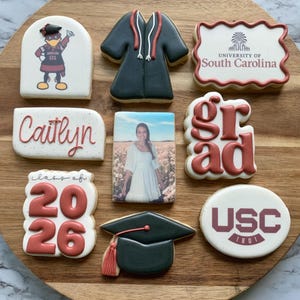 Puede incluir: Una colección de galletas decoradas dispuestas sobre una tabla de madera. Las galletas presentan diseños temáticos de graduación, incluyendo un birrete, una toga de graduación, y los textos "Grad", "Caitlyn", "Class of 2026" y el logo de la Universidad de Carolina del Sur.