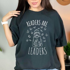 Puede incluir: Camiseta negra con un gráfico blanco de una mujer leyendo un libro con mariposas a su alrededor. El texto "Readers are Leaders" está impreso debajo del gráfico.