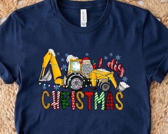 May include: Navy blue t-shirt with a Christmas-themed graphic. The design features a yellow excavator with Santa Claus in the driver's seat, the text "I dig Christmas", and snowflakes. The word "Christmas" is in a colorful, plaid font.