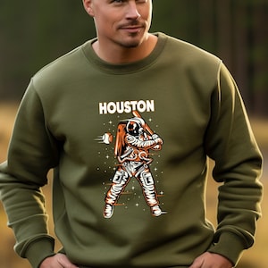 May include: A green sweatshirt with a graphic of an astronaut playing baseball. The text "HOUSTON" is printed above the graphic.