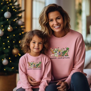 Pu&ograve; includere: Un maglione natalizio rosa con un'immagine di personaggi di Toy Story in cartone animato che indossano cappelli di Babbo Natale. Il testo "Merry Christmas" &egrave; stampato sotto l'immagine. Una donna e una ragazza indossano i maglioni.