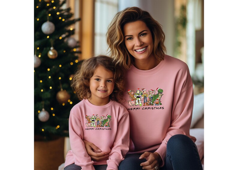 Pu&ograve; includere: Un maglione natalizio rosa con un'immagine di personaggi di Toy Story in cartone animato che indossano cappelli di Babbo Natale. Il testo "Merry Christmas" &egrave; stampato sotto l'immagine. Una donna e una ragazza indossano i maglioni.