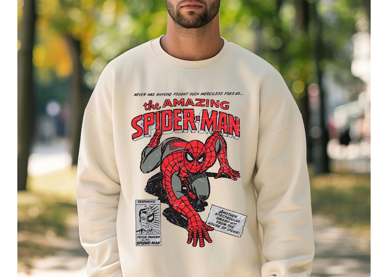 May include: A cream colored sweatshirt with a vintage comic book design featuring Spider-Man. The text reads "Never has anyone fought such merciless foes as... the Amazing Spider-Man".