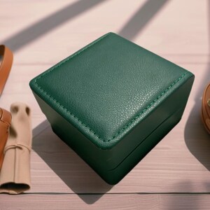May include: A dark green, square-shaped box with a leather-like texture and stitched edges. The box is made of a sturdy material and has a smooth, polished finish.