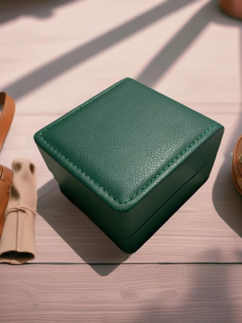 May include: A dark green, square-shaped box with a leather-like texture and stitched edges. The box is made of a sturdy material and has a smooth, polished finish.