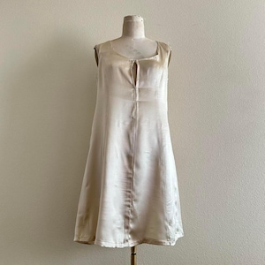 May include: A sleeveless, knee-length dress in a light beige color. The dress has a scoop neckline with a keyhole detail and a subtle sheen, suggesting a satin or silk fabric. It is displayed on a mannequin.