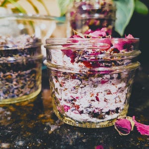 May include: Three small glass jars filled with dried flowers and herbs. The jars contain a mix of pink rose petals, lavender, and other botanicals. The jars are on a dark, speckled surface, with greenery in the background.