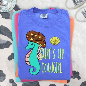 May include: A blue short-sleeved t-shirt featuring a graphic of a teal seahorse wearing a brown cowboy hat with yellow hearts. Next to the seahorse is a yellow seashell and the text Surf's Up Cowgirl in yellow lettering. The shirt has a tag.