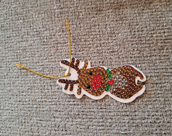 May include: A festive reindeer Christmas ornament. The reindeer is adorned with brown and clear rhinestones, a red nose, and a green collar. It has a gold hanging string.