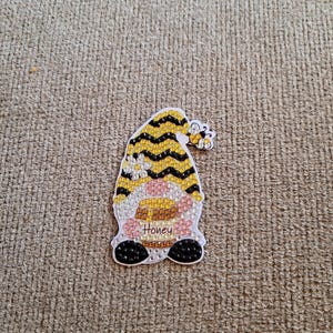 Peut inclure: Un gnome rayé jaune et noir avec une abeille sur son chapeau. Le gnome tient un rayon de miel et a le mot "Honey" écrit sur son ventre.