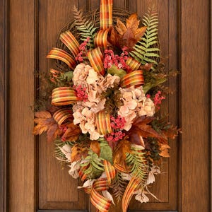 May include: A teardrop-shaped autumn wreath featuring a grapevine base, light pink hydrangea blossoms, and brown maple leaves. It is accented with green fern fronds, small red berries, and a plaid ribbon in shades of orange, yellow, and green.