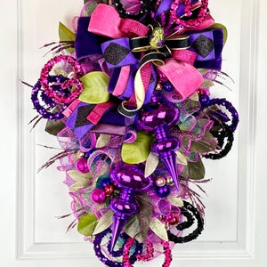 May include: A purple, pink, and black Christmas wreath with a large bow and ornaments. The wreath is decorated with sequins and mesh.