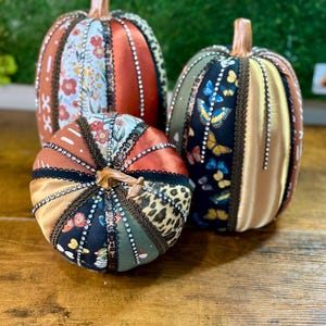 May include: Three decorative fabric pumpkins in various sizes. The pumpkins feature a mix of patterns, including floral, leopard print, and butterfly designs. They are adorned with brown trim and small embellishments, with a natural-looking stem.