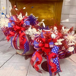 May include: Patriotic floral arrangements in decorative containers. The arrangements feature red, white, and blue ribbons, stars, and decorative elements. The containers are in a distressed finish, adding a vintage touch to the festive decor.
